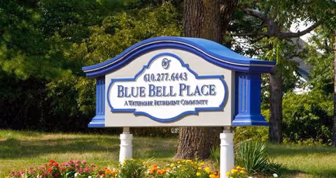 A decorative outdoor sign for Blue Bell Place, a Watermark Retirement Community, with a blue and white color scheme, set in a landscaped area with flowers and trees in the background.