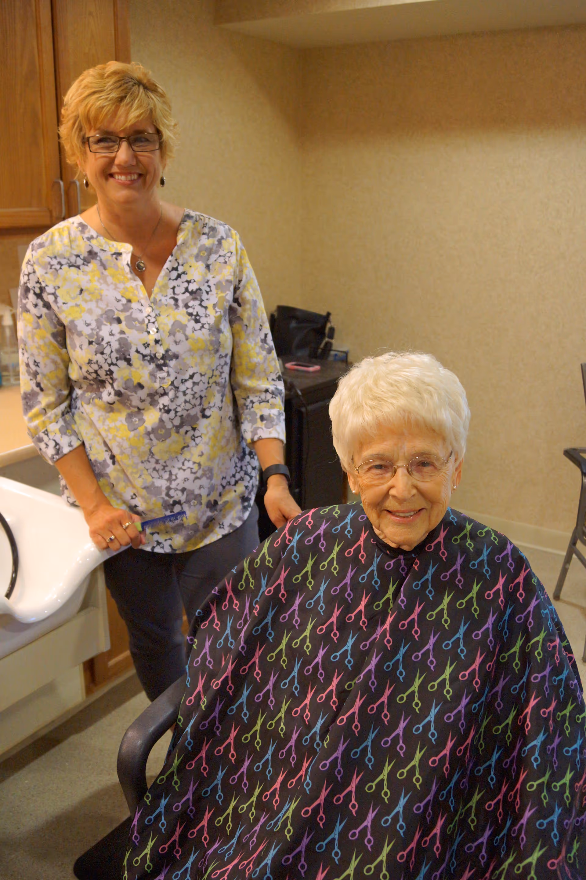 An elderly woman with white hair and glasses sits in a chair wearing a black haircut cape with colorful scissors pattern. A smiling woman with short blonde hair, glasses, and a floral blouse stands behind her in a room with beige walls and wooden cabinets, likely a salon or hair care area in a senior living facility.