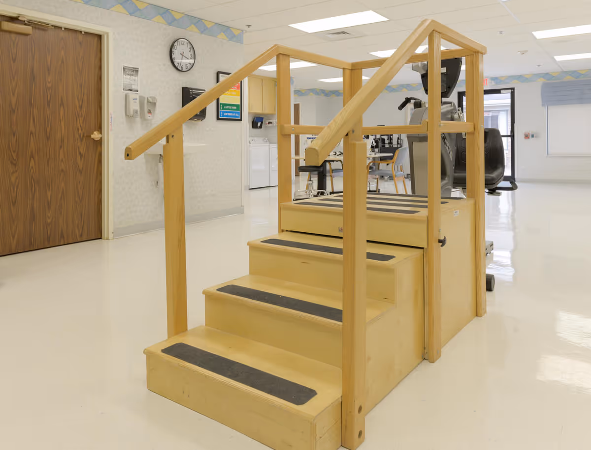 Indoor physical therapy area with a wooden exercise staircase with handrails, exercise equipment, chairs, and a clock on the wall in a well-lit room.