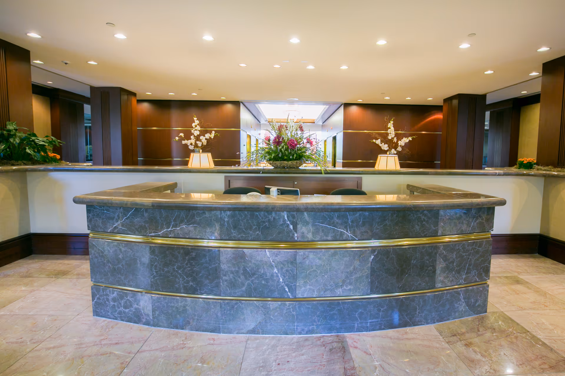A spacious reception desk area with a curved marble counter featuring gold accents. Behind the desk are two chairs, a telephone, and a large floral arrangement in the center with two smaller flower arrangements on either side. The background includes wood-paneled walls and a well-lit hallway.