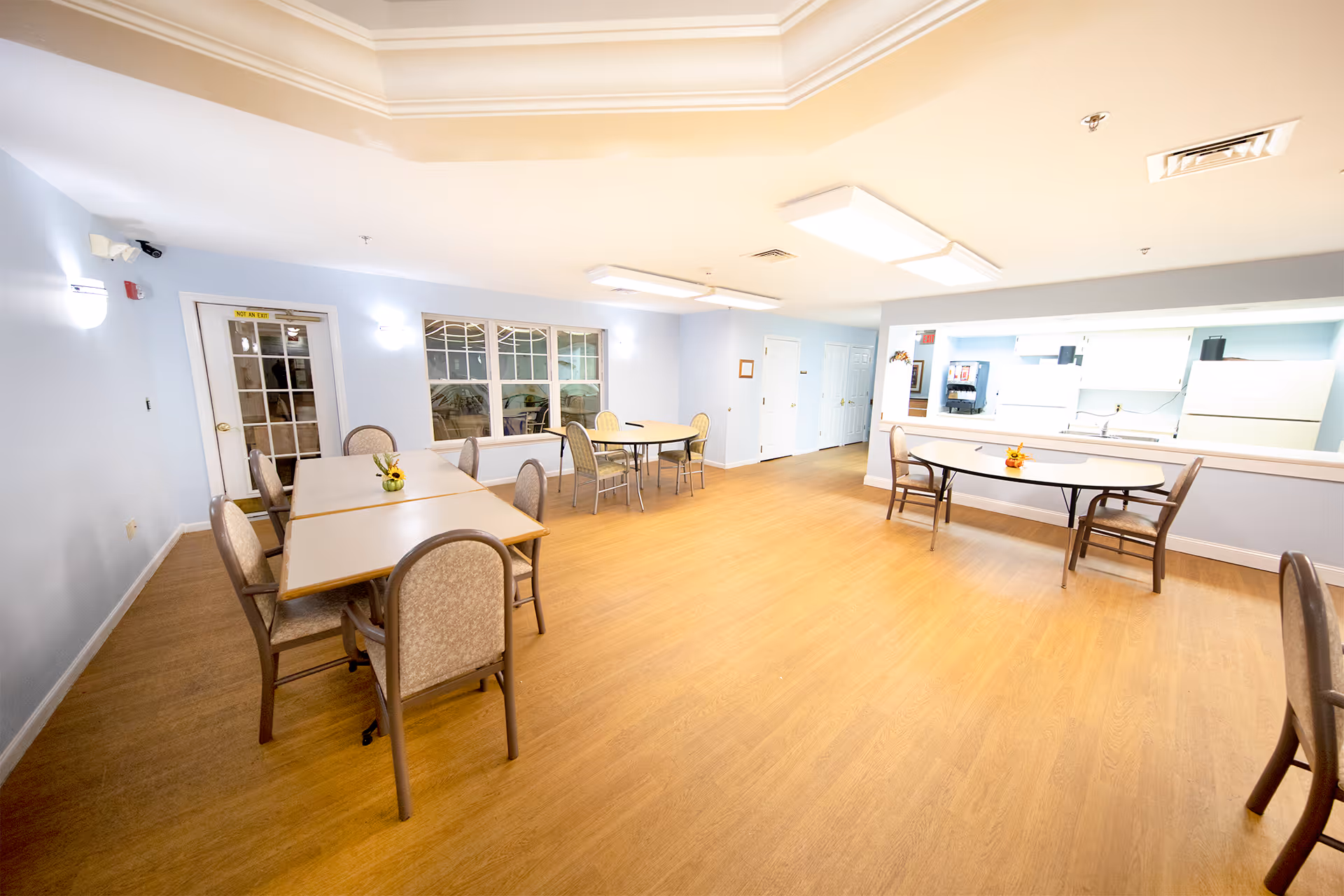 A spacious dining room with light blue walls and wood flooring. The room contains several tables with chairs arranged around them. There are small flower arrangements on the tables. A large window and a door are visible on one wall, and a pass-through window to a kitchen area is on another wall. The room is well-lit with ceiling lights.