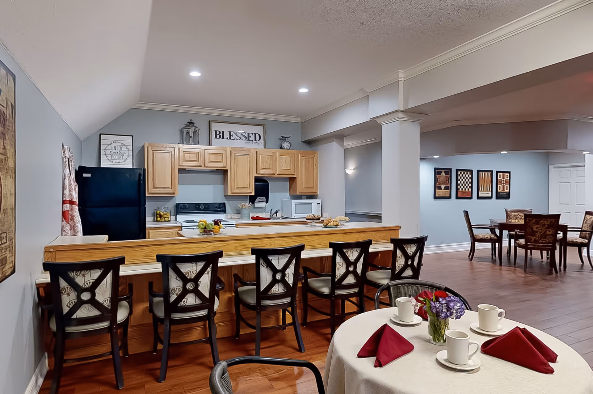 Interior view of a communal kitchen and dining area in an assisted living facility. The kitchen features wooden cabinets, a black refrigerator, a stove, microwave, and a counter with six high chairs. In the foreground, there is a round table set with white cups, saucers, red napkins, and a small flower arrangement. In the background, there are additional dining tables and chairs against light blue walls decorated with framed artwork.