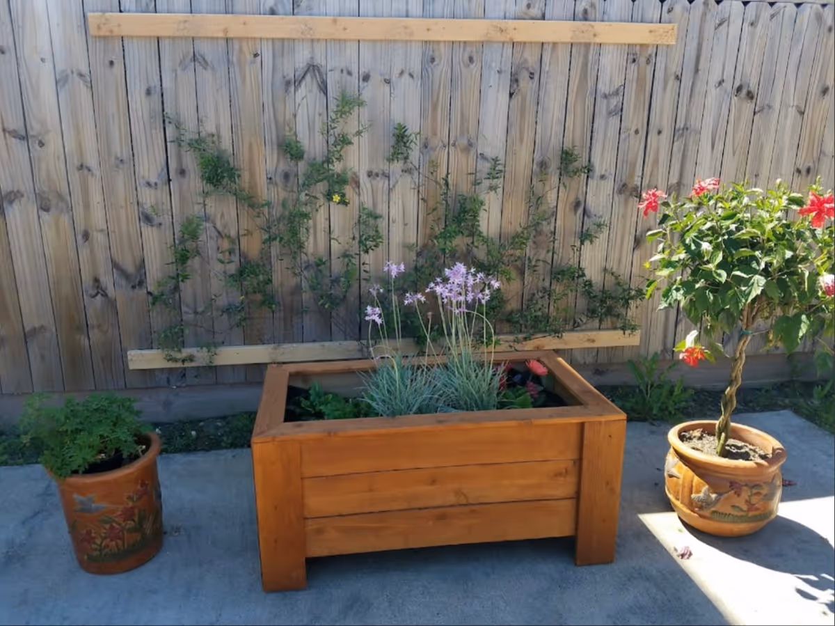 Wooden planter box with flowering plants and two decorative pots on a concrete patio in front of a wooden fence.