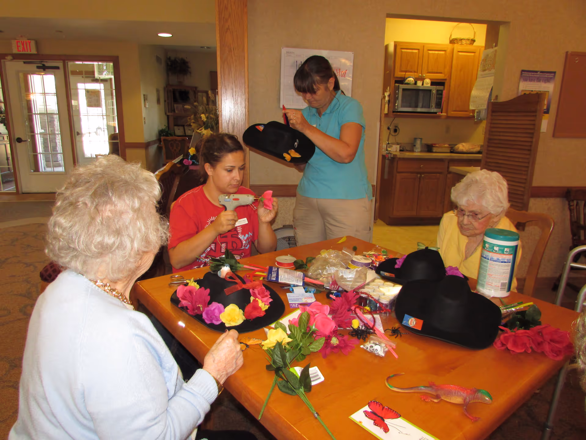 Four women sitting and standing around a wooden table in a communal room, engaged in a craft activity decorating black hats with colorful artificial flowers and other craft supplies. The room has a kitchen area in the background and an exit door visible to the left.