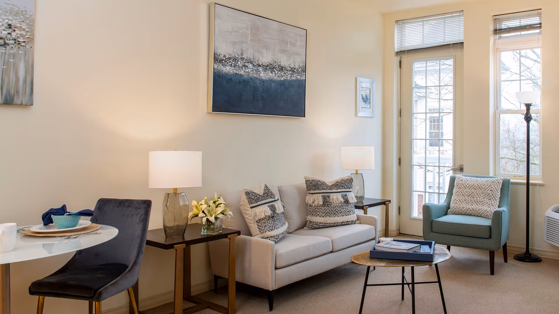A cozy living room with a beige sofa adorned with patterned cushions, a blue armchair with a decorative pillow, a round wooden coffee table with books, two side tables each with a lamp, a small round dining table with a chair, and large windows letting in natural light.