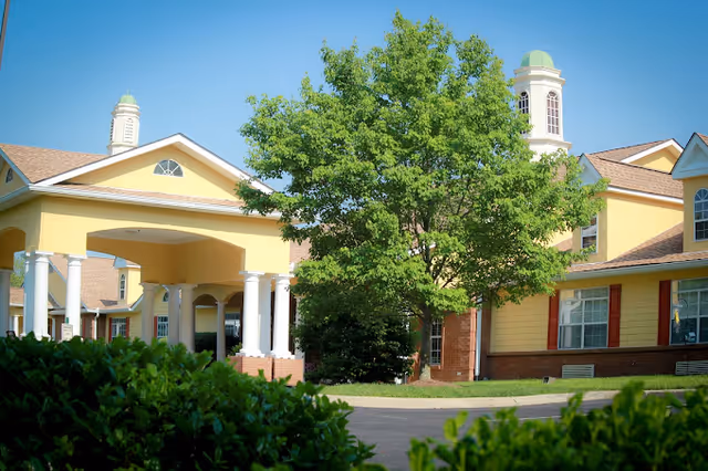 Front entrance of a yellow senior living facility with white columns, a drive, and landscaping including a large tree and shrubs.