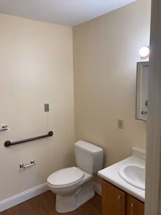 A small bathroom with beige walls, a white toilet, a white sink with a wooden cabinet underneath, a mirror above the sink, a black grab bar on the wall next to the toilet, and a toilet paper holder without a roll.