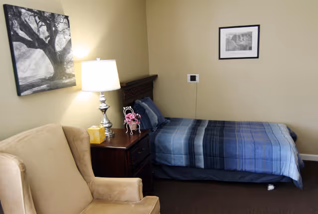 A small bedroom with a single bed covered in a blue striped blanket, a wooden nightstand with a lamp, tissue box, and decorative items, a beige armchair, and two black and white framed pictures on the walls.