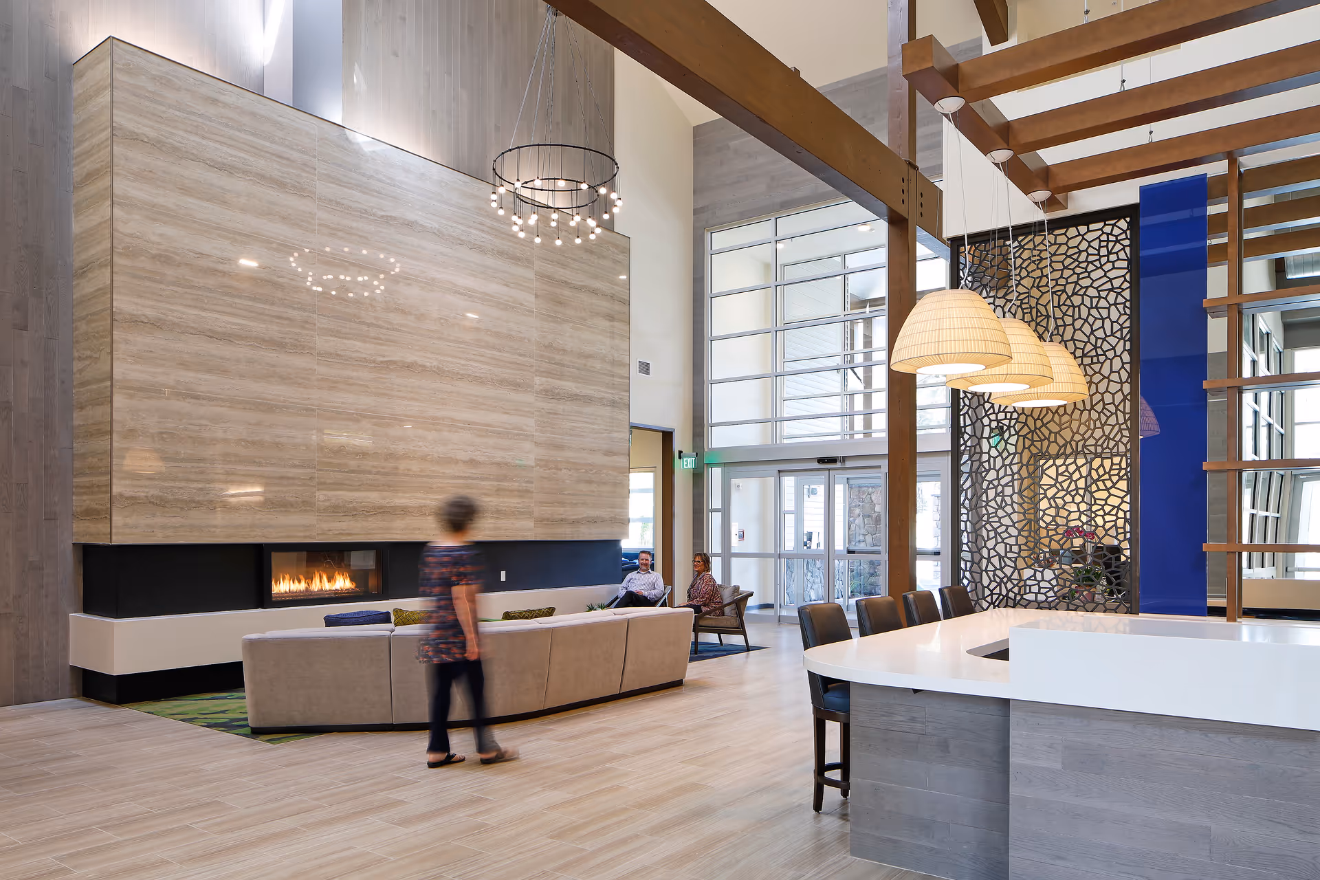 A modern, spacious living room area in a senior living facility with a large beige sectional sofa facing a built-in fireplace set in a tall, light-colored stone wall. A woman is walking past the sofa, and two people are seated on chairs near the glass entrance doors. The room features high ceilings with wooden beams, a circular chandelier, and pendant lights hanging over a white countertop bar area.