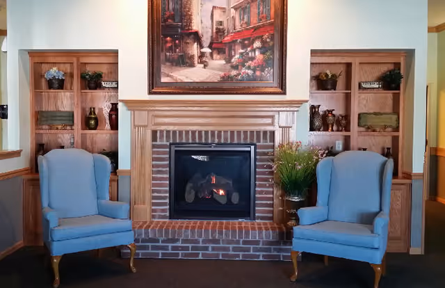 A cozy interior living area featuring a brick fireplace with a wooden mantle. Above the fireplace hangs a framed painting of a street scene with flowers and buildings. On either side of the fireplace are built-in wooden shelves decorated with vases, plants, and small signs. Two light blue upholstered armchairs with wooden legs are positioned facing the fireplace.
