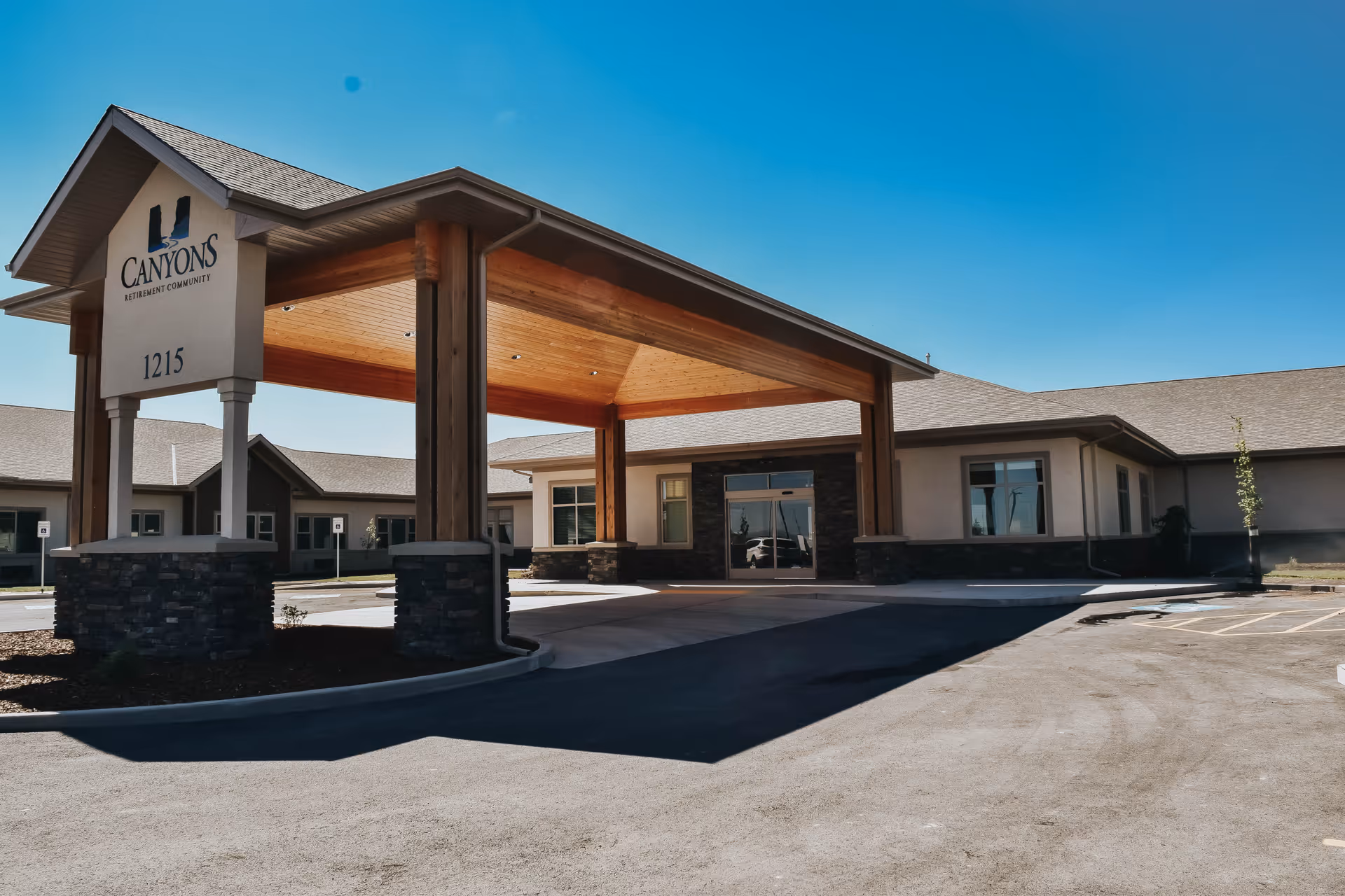 Exterior view of Canyons Senior Living retirement community building with a covered entrance supported by wooden pillars and stone bases, clear blue sky in the background.