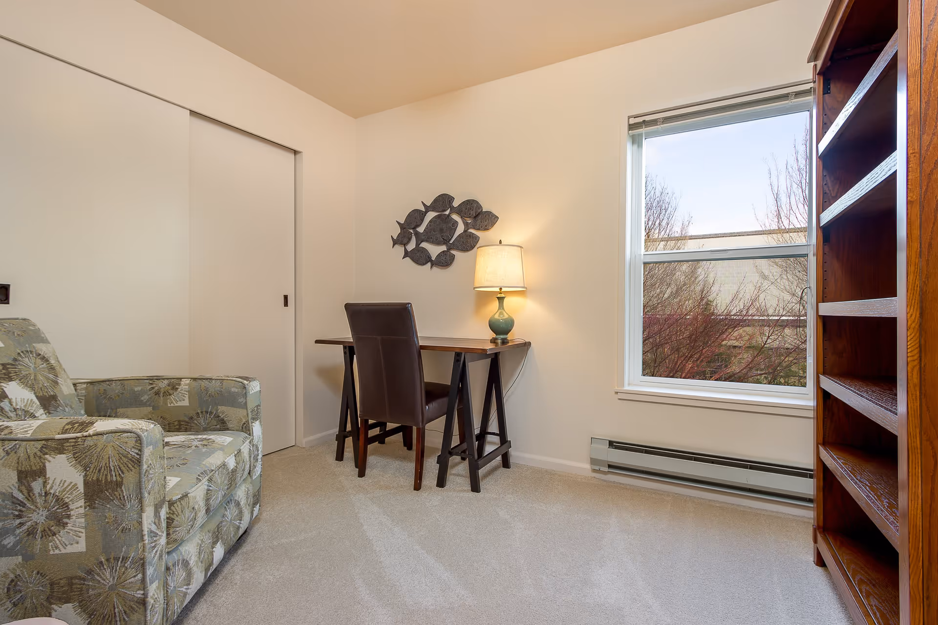 Small furnished living room/office with an armchair, desk and chair, lamp, bookshelf, and a window.