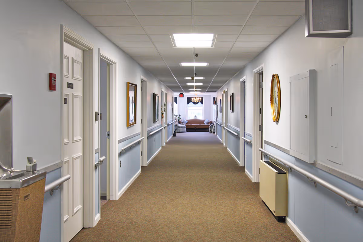 Long, well-lit interior hallway of a senior living facility with doors and handrails leading to a seating area at the far end.