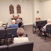 A group of elderly people seated in chairs facing a woman who appears to be reading or speaking in a room with beige walls and carpeted floor. There are decorative stained glass window panels on the wall behind the woman and a small table with a framed picture on the right side. Some walkers are visible near the seated individuals.