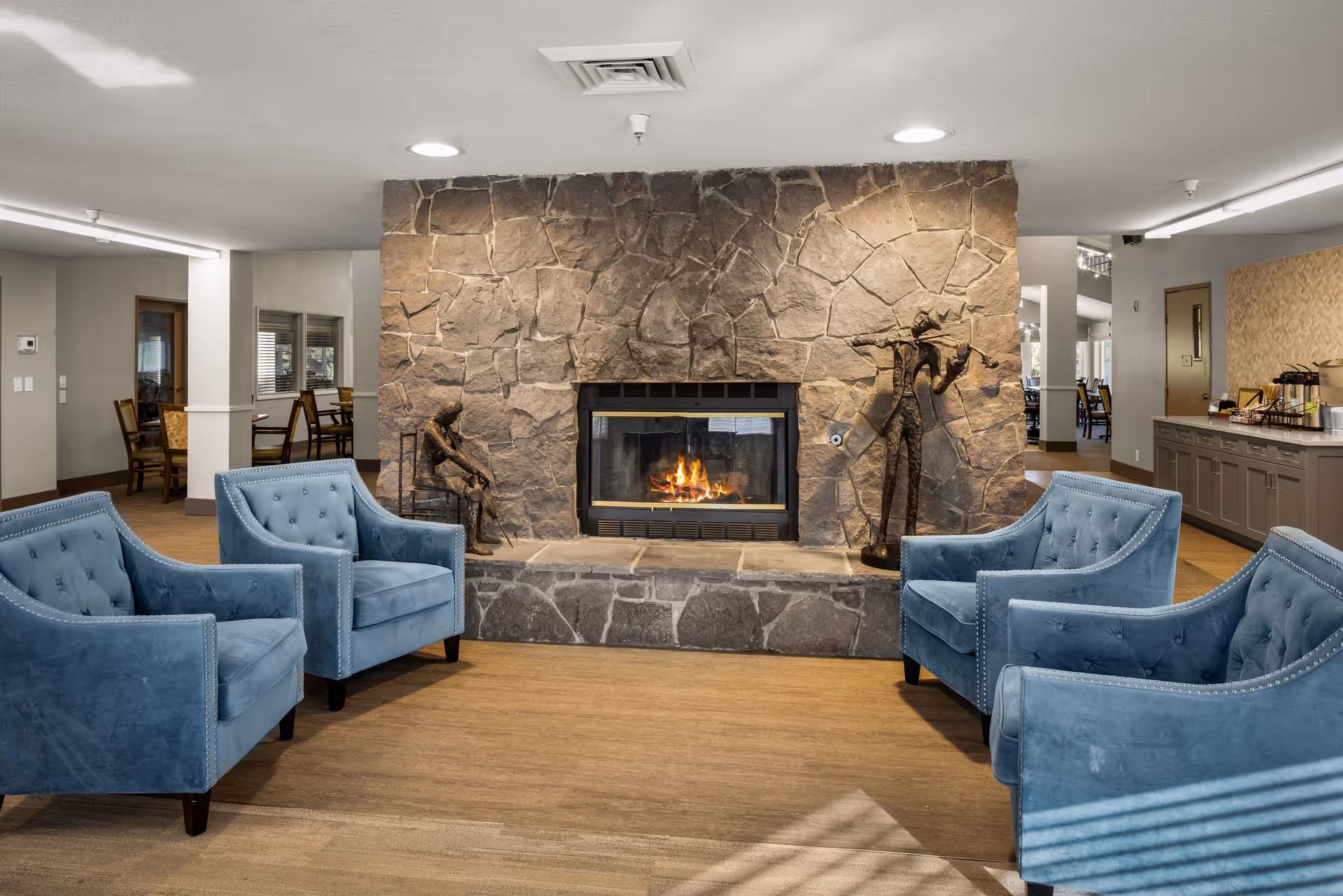 A cozy seating area in a senior living community featuring four blue upholstered armchairs arranged around a stone fireplace with a fire burning. Two artistic metal sculptures are placed on either side of the fireplace. The room has wood flooring and neutral-colored walls, with a dining area visible in the background.