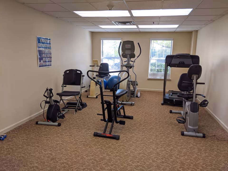 A small exercise room with various fitness equipment including stationary bikes, an elliptical machine, a treadmill, and exercise balls. The room has beige walls, carpeted floor, two windows letting in natural light, and a poster on the left wall.