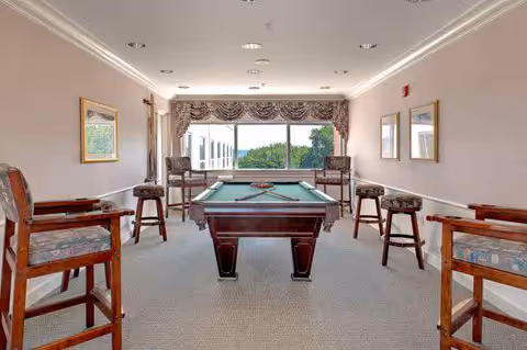A well-lit game room with a pool table in the center, surrounded by six wooden chairs with patterned cushions. The room has beige walls, a large window with a decorative valance, and framed pictures on the walls.