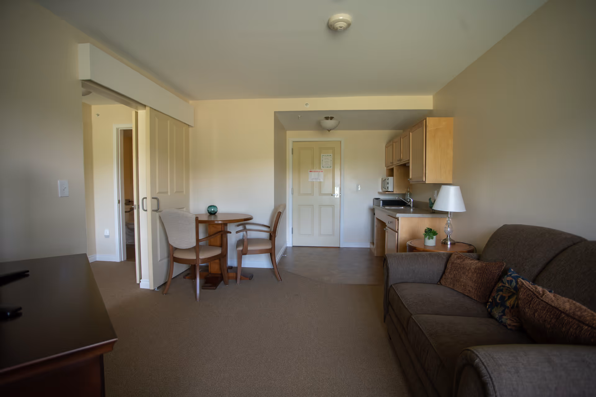 Interior view of a small assisted living apartment featuring a brown sofa with decorative pillows on the right, a round wooden table with two chairs in the center, a kitchenette with wooden cabinets, microwave, and sink on the right side near the entrance door, and a sliding door leading to another room on the left.