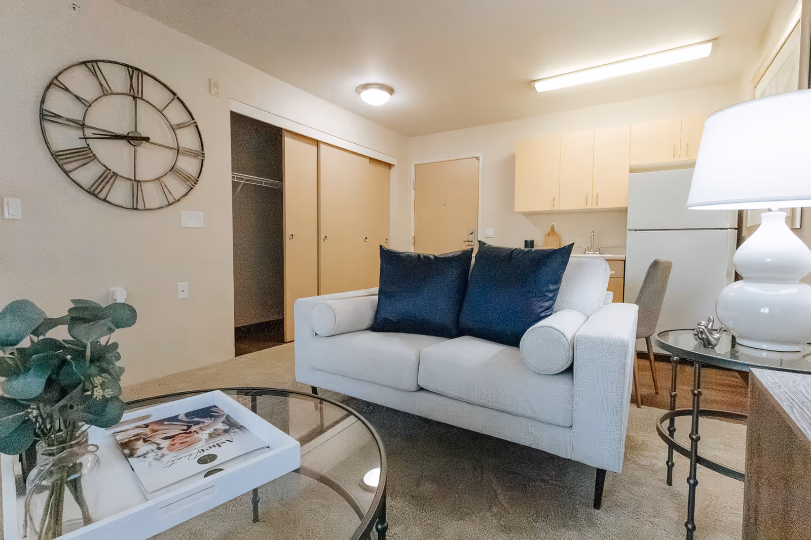 Small living area featuring a white loveseat with navy pillows, glass coffee table, lamp and a kitchenette with refrigerator in the background.
