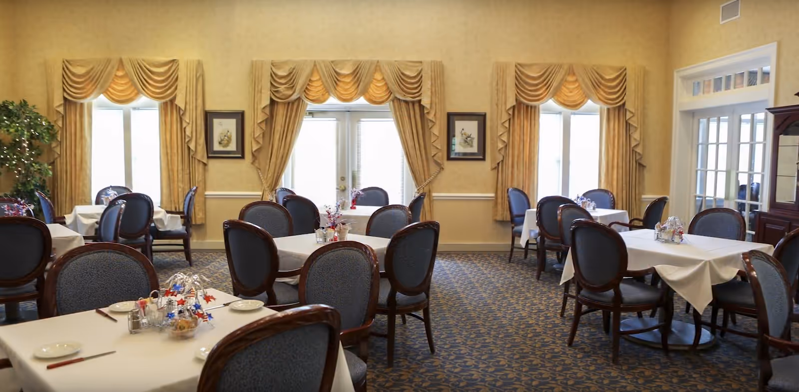 A dining room with multiple tables covered in white tablecloths, each surrounded by blue upholstered chairs with wooden frames. The room has patterned carpet, large windows with gold drapes, and framed artwork on the walls. There are decorative centerpieces on the tables and a glass cabinet on the right side.