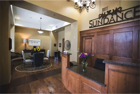 Warmly lit reception desk and adjoining seating area in a senior living facility lobby with wood finishes and a 'SUNDANCE' sign.