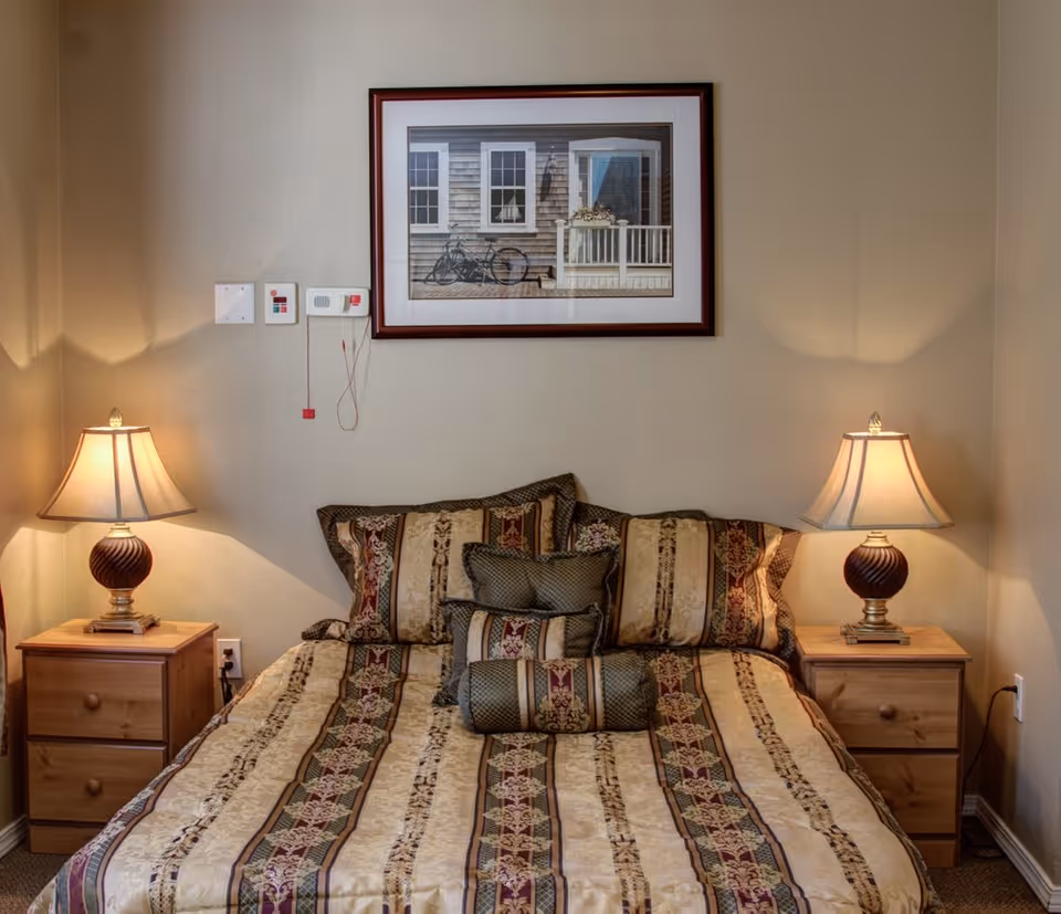 A neatly made bed with patterned bedding and multiple pillows is centered between two wooden nightstands, each with a lamp. Above the bed hangs a framed picture of a porch with a bicycle. The walls are painted beige and there are some control panels on the wall to the left of the picture.