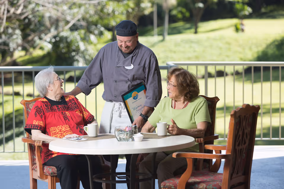Two elderly women sitting at a round outdoor table with coffee mugs, smiling and interacting with a chef who is standing beside them holding a menu. The setting is a sunny patio with a green lawn and trees in the background.
