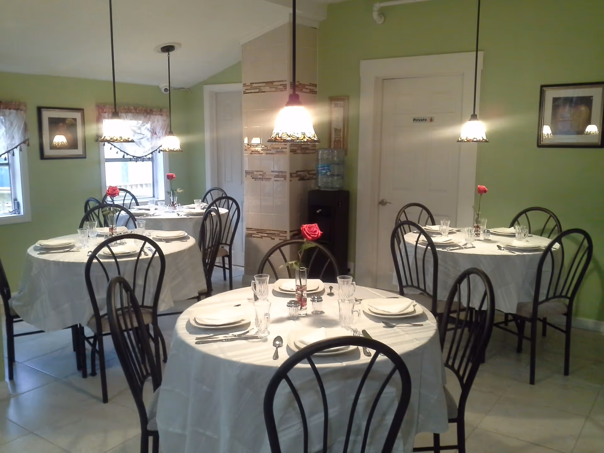 A dining room with three round tables covered with white tablecloths, each set with plates, glasses, silverware, and a single red rose in a vase. The room has light green walls, two windows with sheer curtains, framed pictures on the walls, and three pendant lights hanging from the ceiling. There is a door labeled 'Private' and a water dispenser in the corner.
