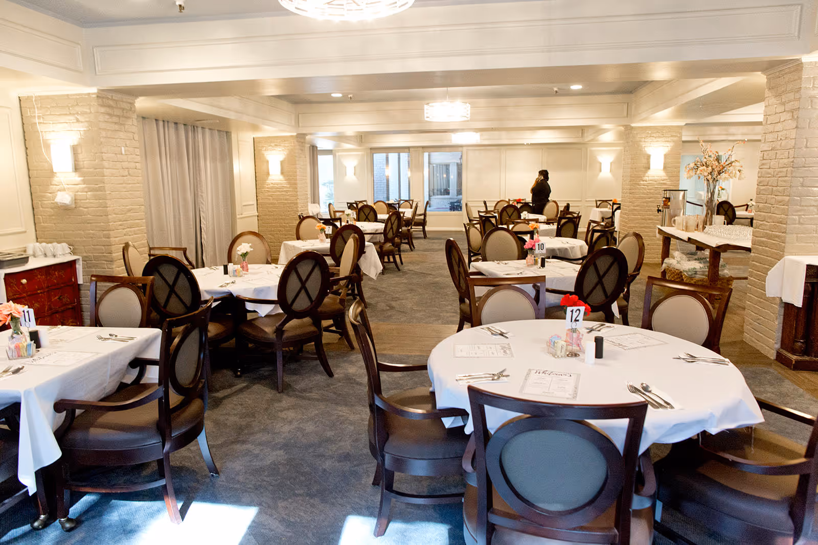 A bright dining room with several round tables covered with white tablecloths, each set with menus, utensils, and small flower arrangements. The room has beige brick pillars, soft lighting, and a carpeted floor. A person is seen standing near the back of the room.