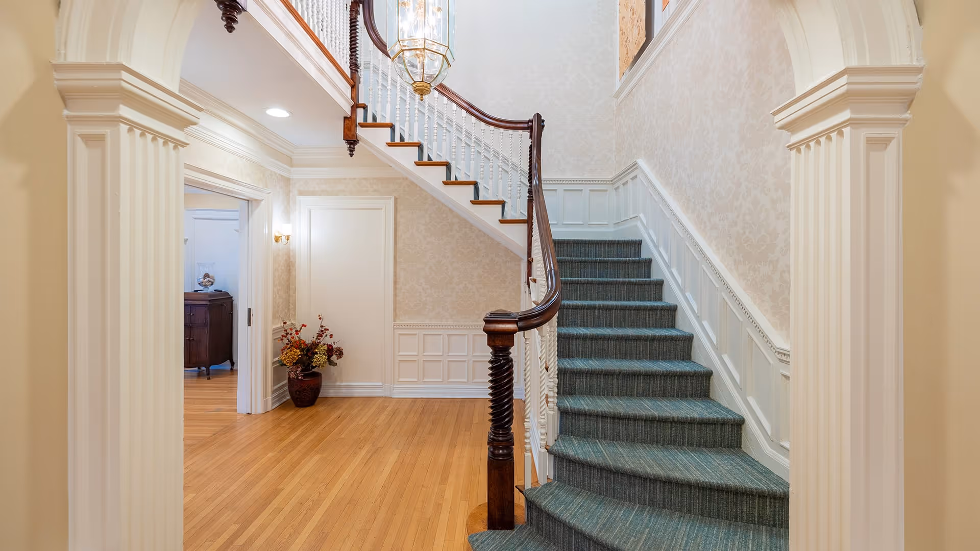 Interior view of a well-lit hallway featuring a wooden staircase with green carpeted steps and a polished wooden handrail. The walls are decorated with light-colored wallpaper and white wainscoting. There is a large hanging light fixture above the staircase and a vase with flowers placed in the corner near a doorway leading to another room with wooden furniture.