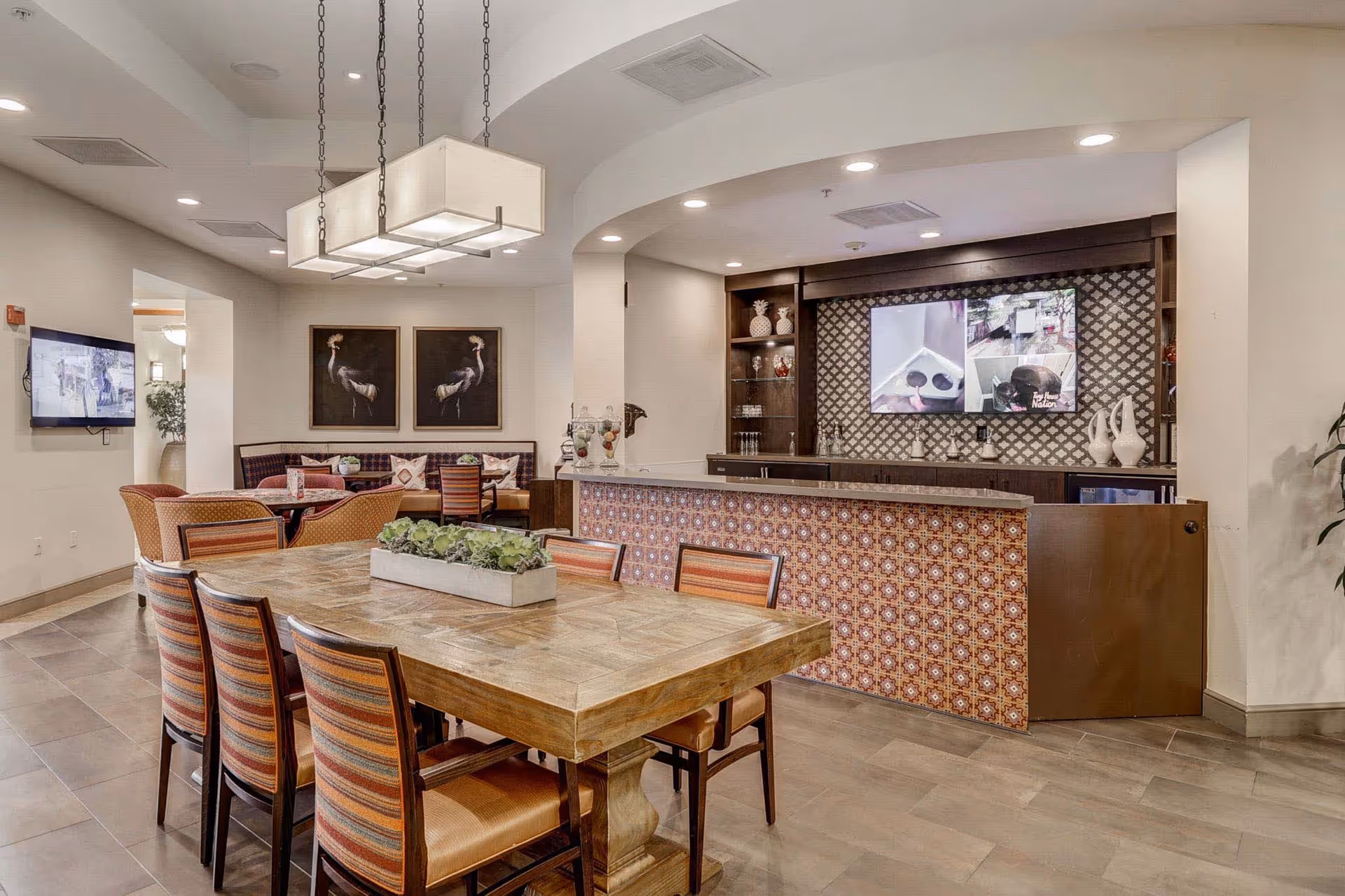 A spacious and well-lit common area in a senior living facility featuring a large wooden table with six striped cushioned chairs and a rectangular planter with green plants in the center. In the background, there is a bar counter with decorative tile facing, shelves with vases and glasses, and a large TV screen mounted on the wall. To the left, there is a smaller seating area with a round table and chairs, and two framed bird artworks on the wall. The room has a modern ceiling light fixture and neutral-colored walls and flooring.