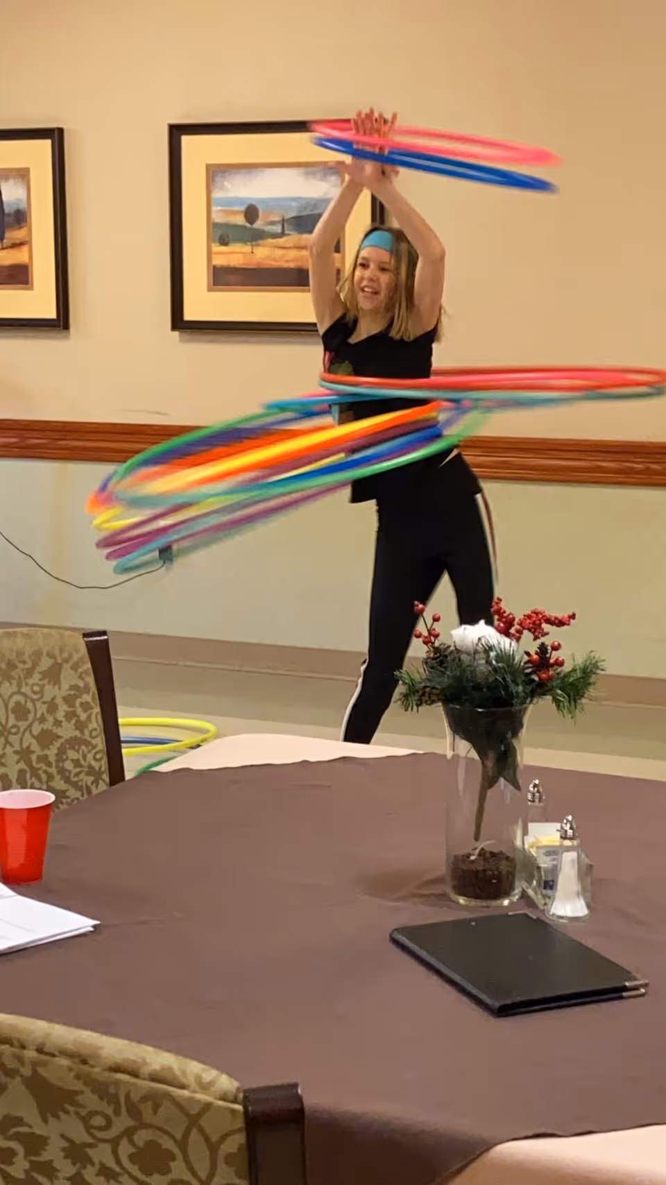 A woman indoors spinning multiple colorful hula hoops around her body and arms. She is smiling and wearing a black outfit with a blue headband. In the foreground, there is a table covered with a brown tablecloth, a vase with a floral arrangement, salt and pepper shakers, a red cup, and a closed black folder. The room has framed landscape paintings on the wall and patterned chairs around the table.