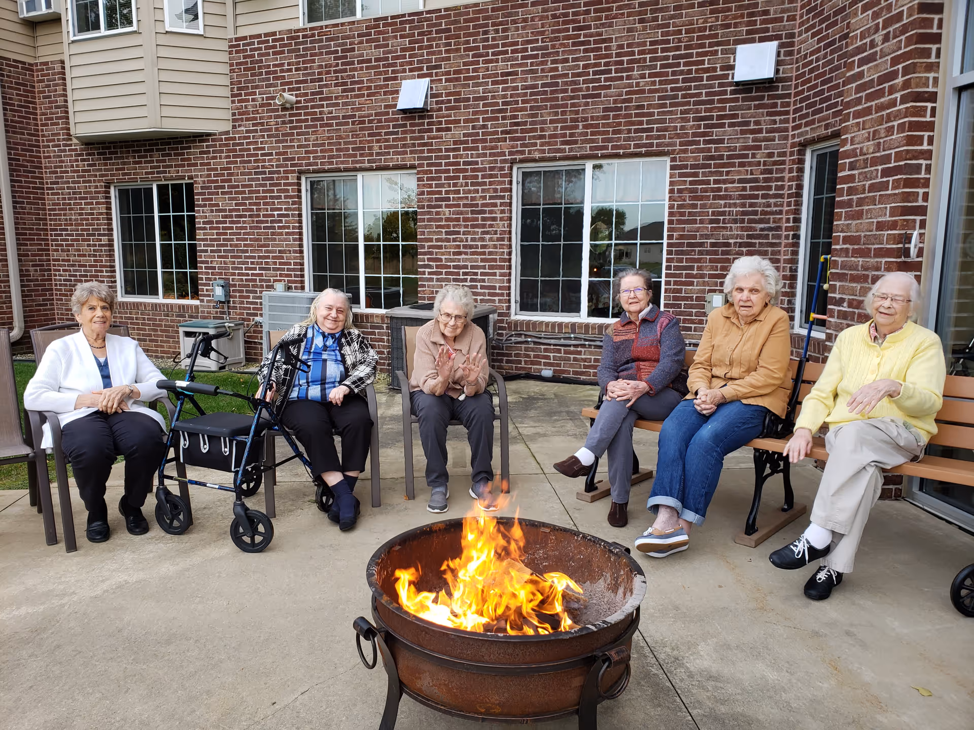 Six elderly women sitting outdoors around a fire pit on a concrete patio in front of a brick building. Some women are seated on chairs and others on a bench, with one walker visible. They appear to be enjoying the warmth of the fire.