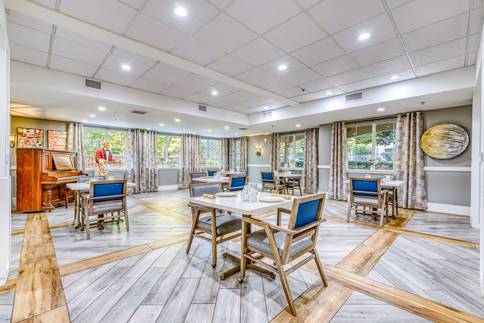A bright and spacious dining room in a senior living facility with several tables and chairs arranged neatly. Large windows with curtains allow natural light to fill the room. A piano and a decorative statue are visible in the corner, and the floor features a patterned wood and tile design.