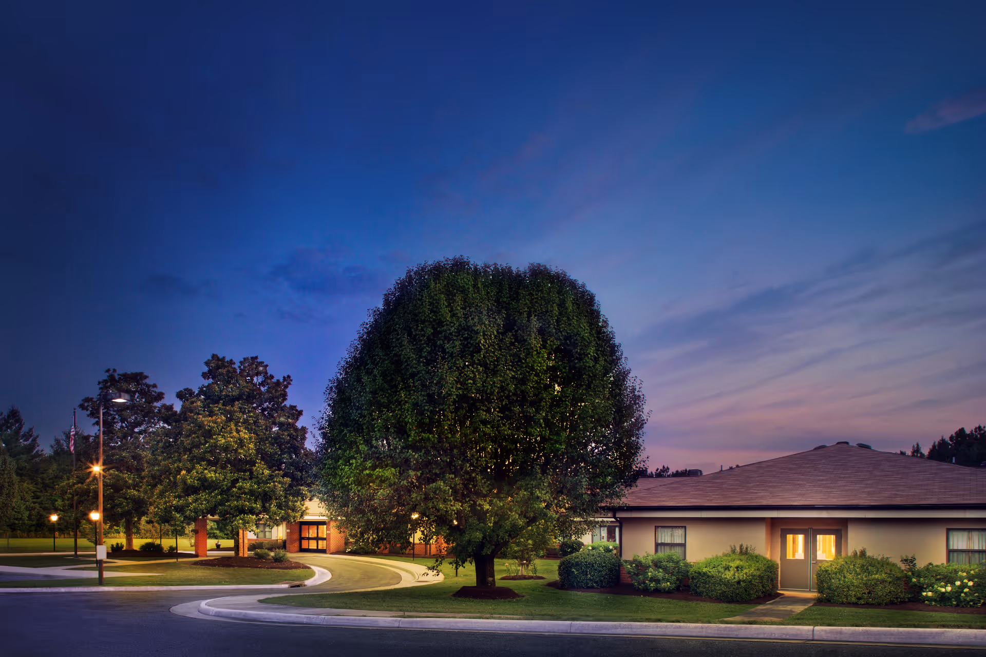 Exterior view of Heritage Hall Brookneal facility at dusk with a large tree in the foreground, well-maintained shrubs, and a curved driveway leading to the entrance of the building.