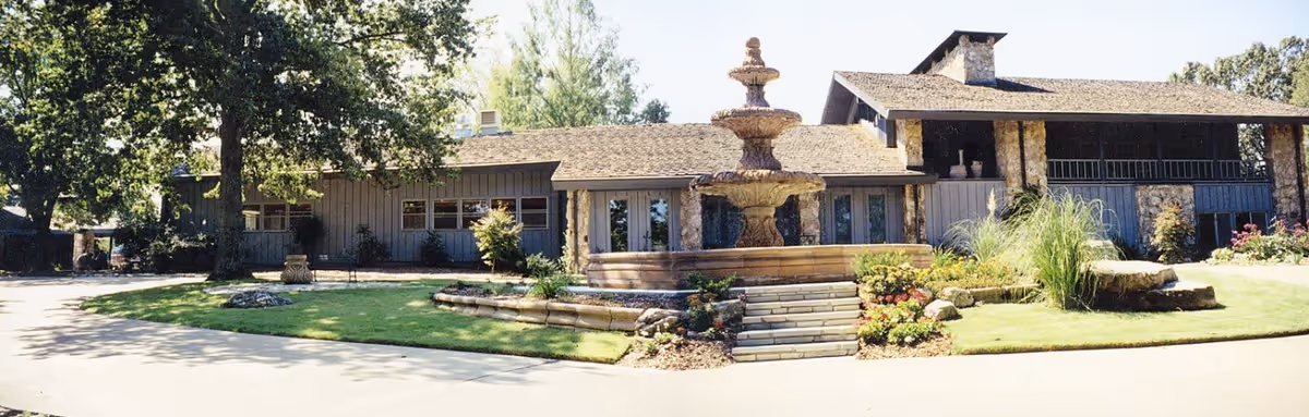 Exterior view of a senior living facility named Fountain Place featuring a large stone fountain in the center of a landscaped circular driveway. The building has a rustic design with stone and wood elements, surrounded by trees and greenery under a clear sky.
