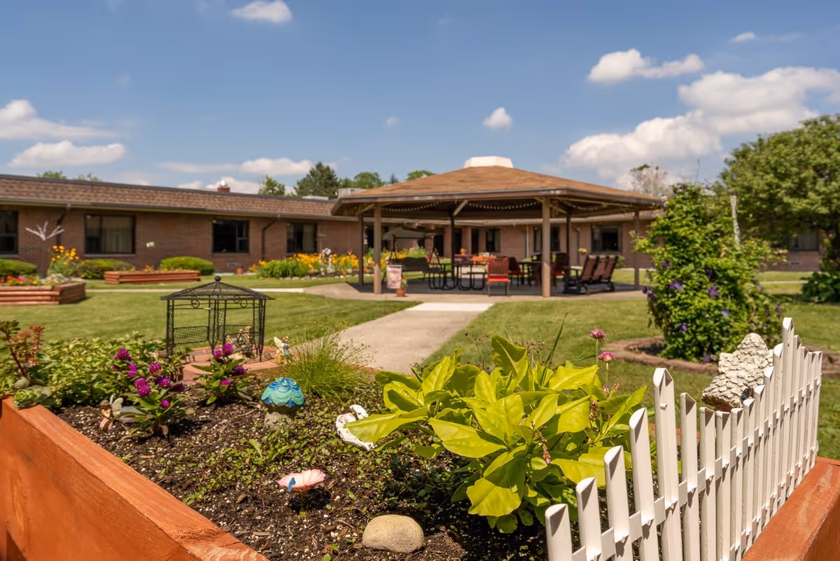 Outdoor garden area with raised flower beds, a small white picket fence, and various plants and flowers. In the background, there is a covered pavilion with seating and tables, surrounded by a single-story brick building under a blue sky with scattered clouds.