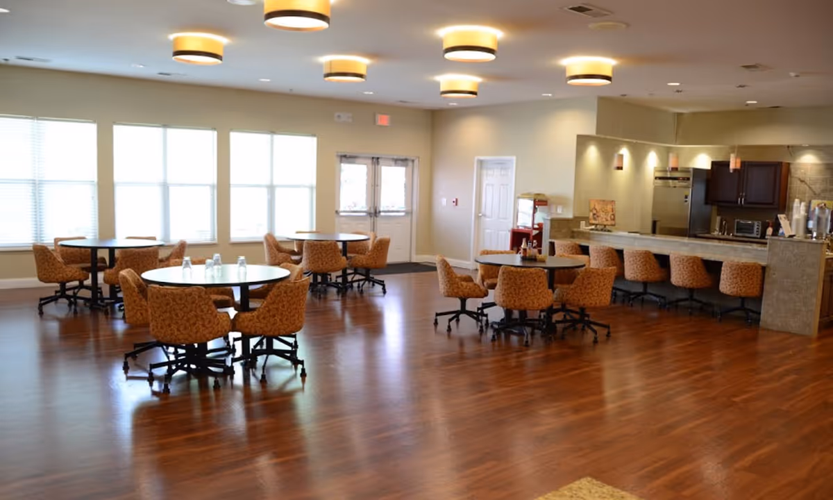 A spacious dining area in a senior living facility with round tables surrounded by cushioned chairs on wheels. The room has large windows letting in natural light, wooden flooring, and a counter area with bar stools and kitchen appliances in the background.