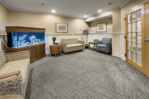 A cozy waiting or sitting area in a senior living facility featuring patterned upholstered chairs and a loveseat, a large built-in aquarium with colorful fish, framed artwork on beige walls, a potted plant, and a wooden door with glass panels.