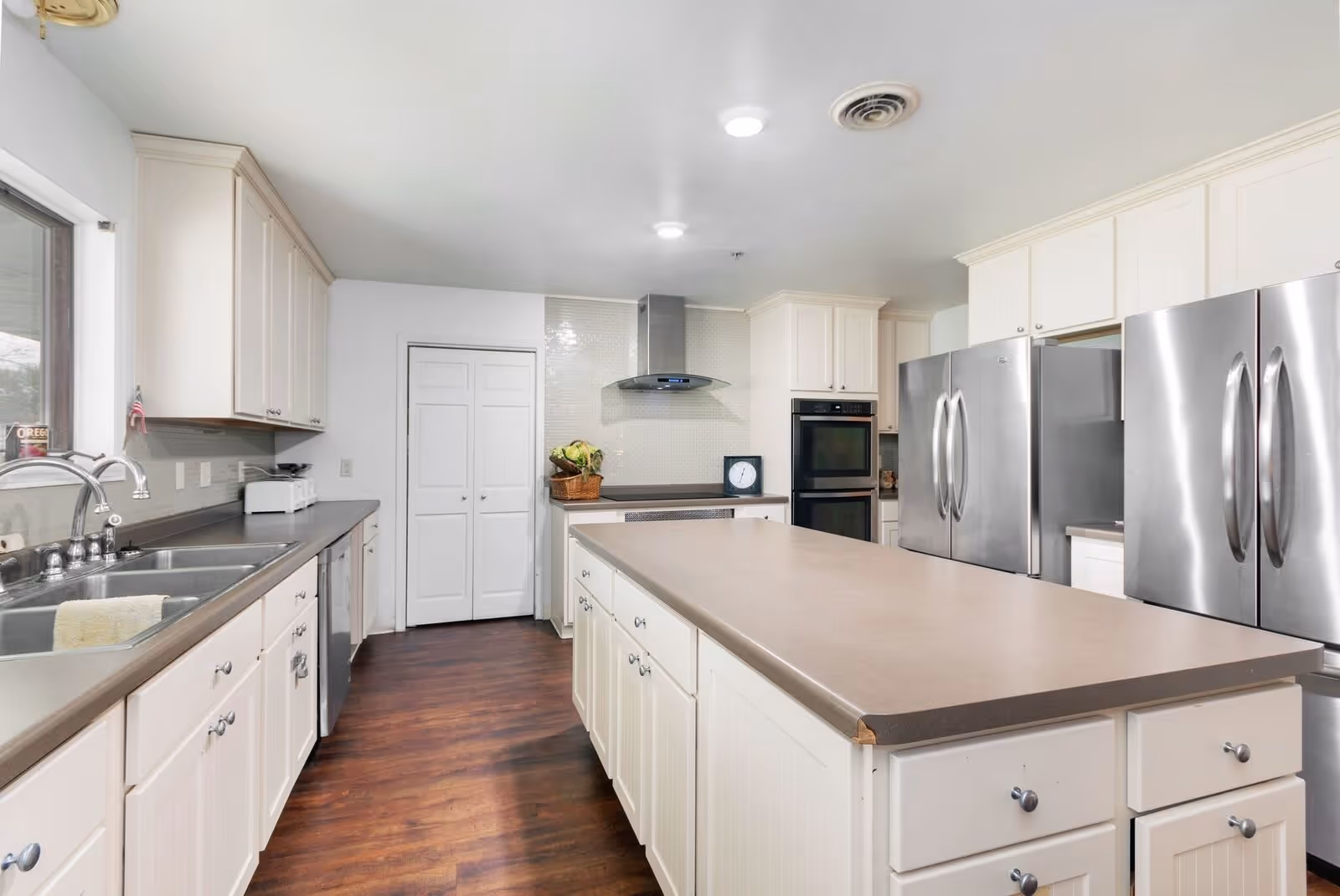 A spacious kitchen with white cabinetry, a large central island with a beige countertop, stainless steel double-door refrigerators, a double wall oven, a stainless steel range hood, and a double sink under a window. The floor is dark wood, and the walls are white with a tiled backsplash behind the stove area.