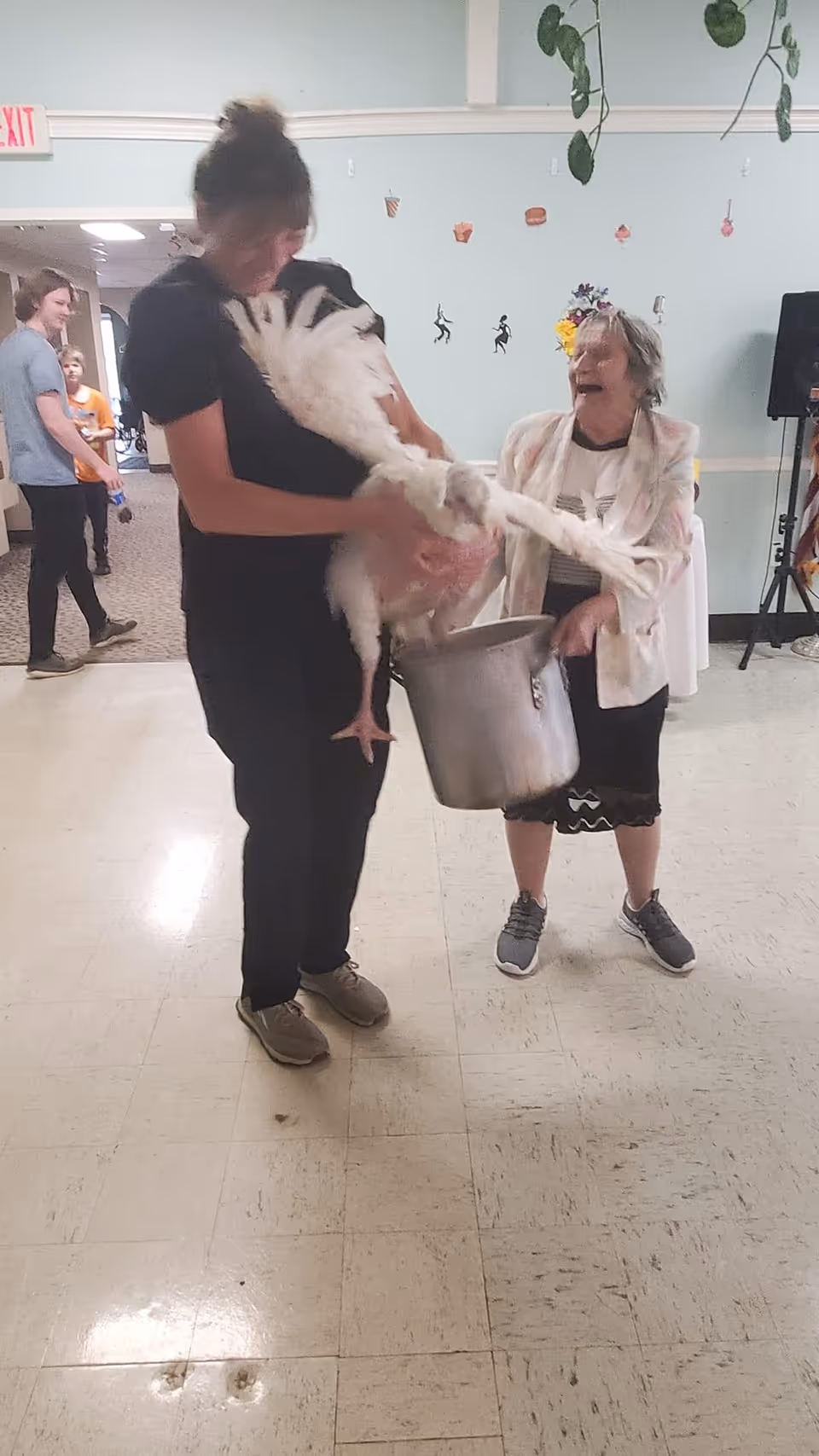 A woman holding a large white bird, possibly a turkey, while an elderly woman stands nearby holding a large metal pot. Both appear to be indoors in a communal area with other people in the background. The elderly woman is smiling and wearing a light-colored jacket and sneakers.