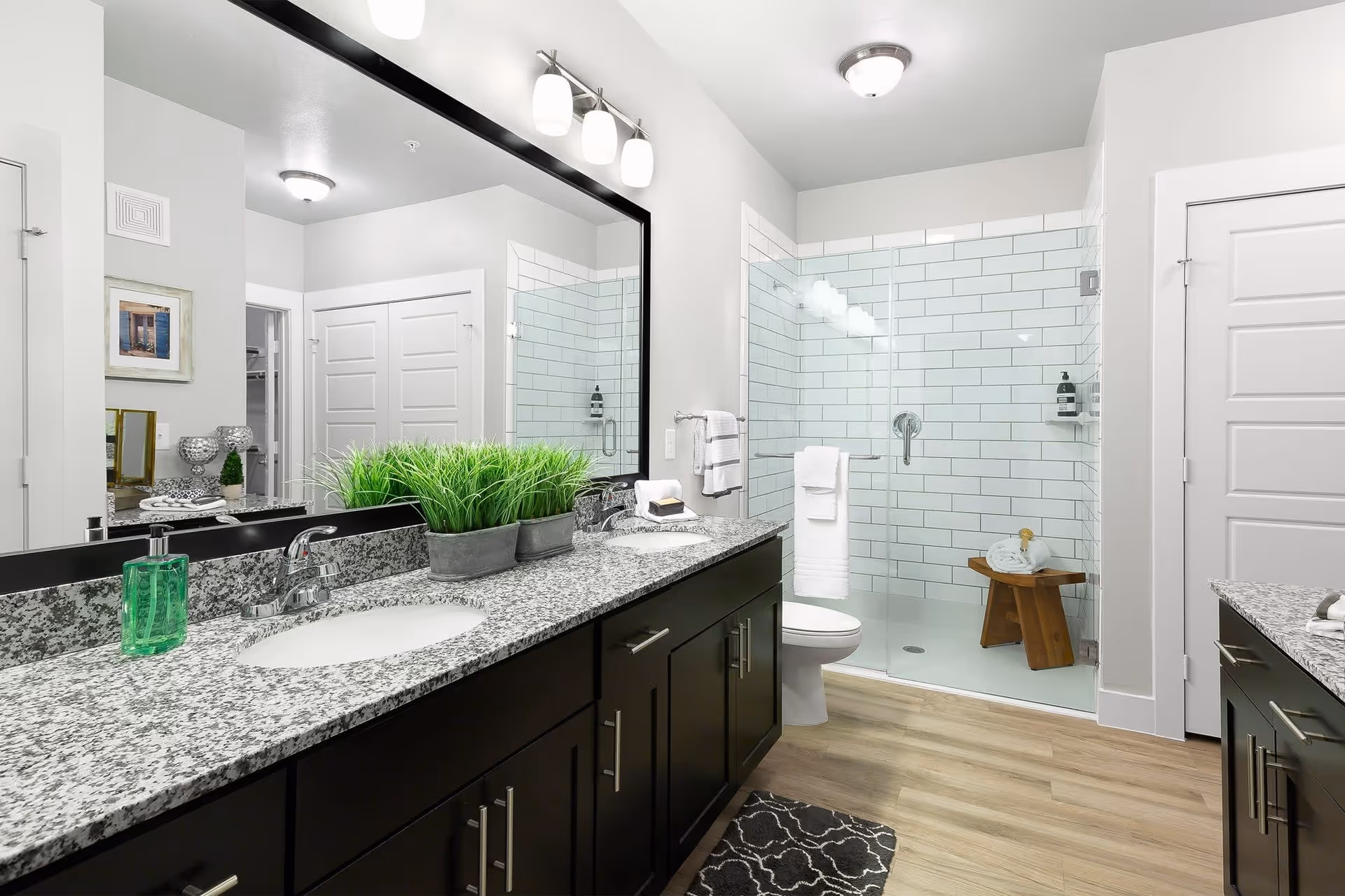 Modern bathroom with a large granite countertop featuring two sinks, a large mirror with three light fixtures above, a glass-enclosed shower with white subway tiles, a wooden stool inside the shower holding rolled towels, a toilet, and light wood flooring.
