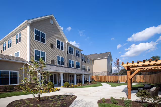 Three-story beige senior living building with a paved courtyard, pergola, and landscaped grounds under a blue sky.