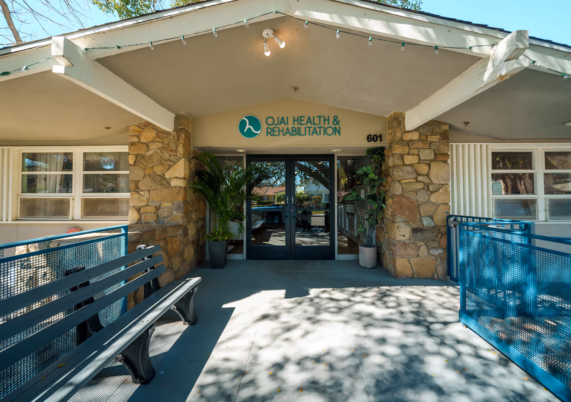 Entrance to Ojai Health & Rehabilitation facility with double glass doors, stone pillars on either side, potted plants near the doors, a bench on the left, and a blue railing on the right under a covered walkway.