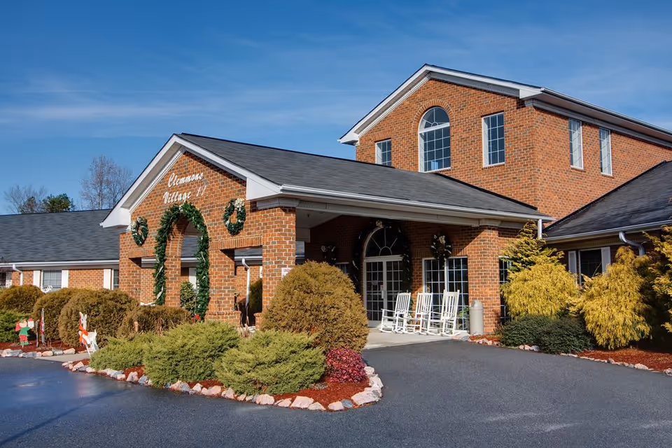 Exterior view of Clemmons Village II Assisted Living facility, a brick building with a covered entrance decorated with green wreaths. There are white rocking chairs on the porch and landscaped bushes and shrubs around the driveway.
