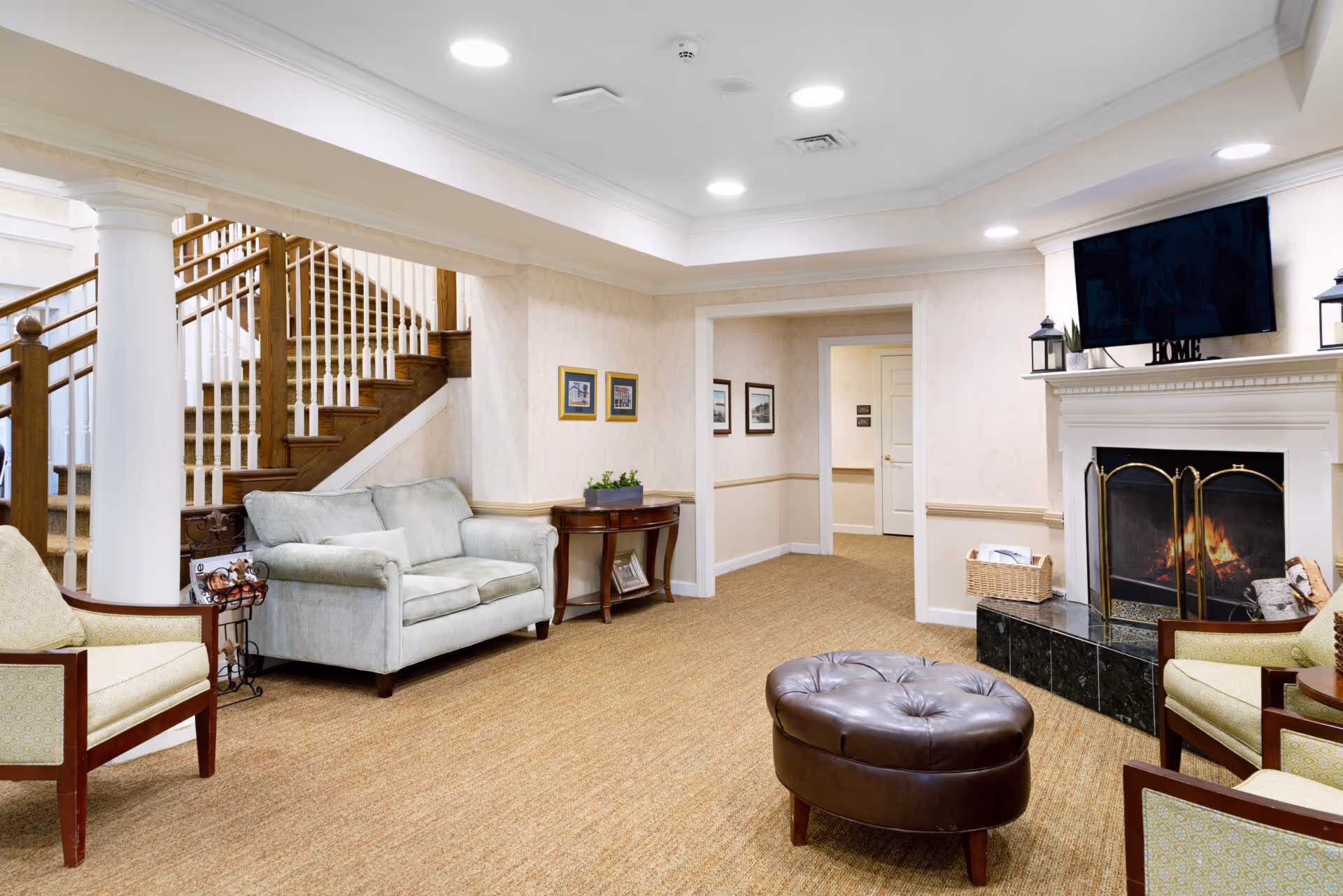 A cozy living room area with beige carpet, light-colored walls, and recessed ceiling lights. The room features a light blue loveseat, two armchairs with wooden frames, a round brown leather ottoman, and a white fireplace with a fire burning inside. Above the fireplace is a mounted flat-screen TV and decorative items including lanterns and a 'HOME' sign. A staircase with wooden handrails and white balusters is visible in the background, along with a small wooden table with plants and framed pictures on the walls.