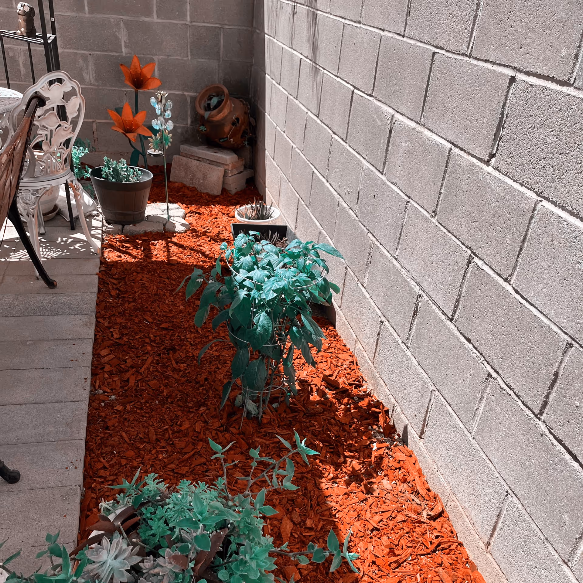 A small outdoor garden area with red mulch covering the ground, several potted plants, and decorative metal flowers. There is a concrete block wall on the right side and a white metal chair and table set on the left side.