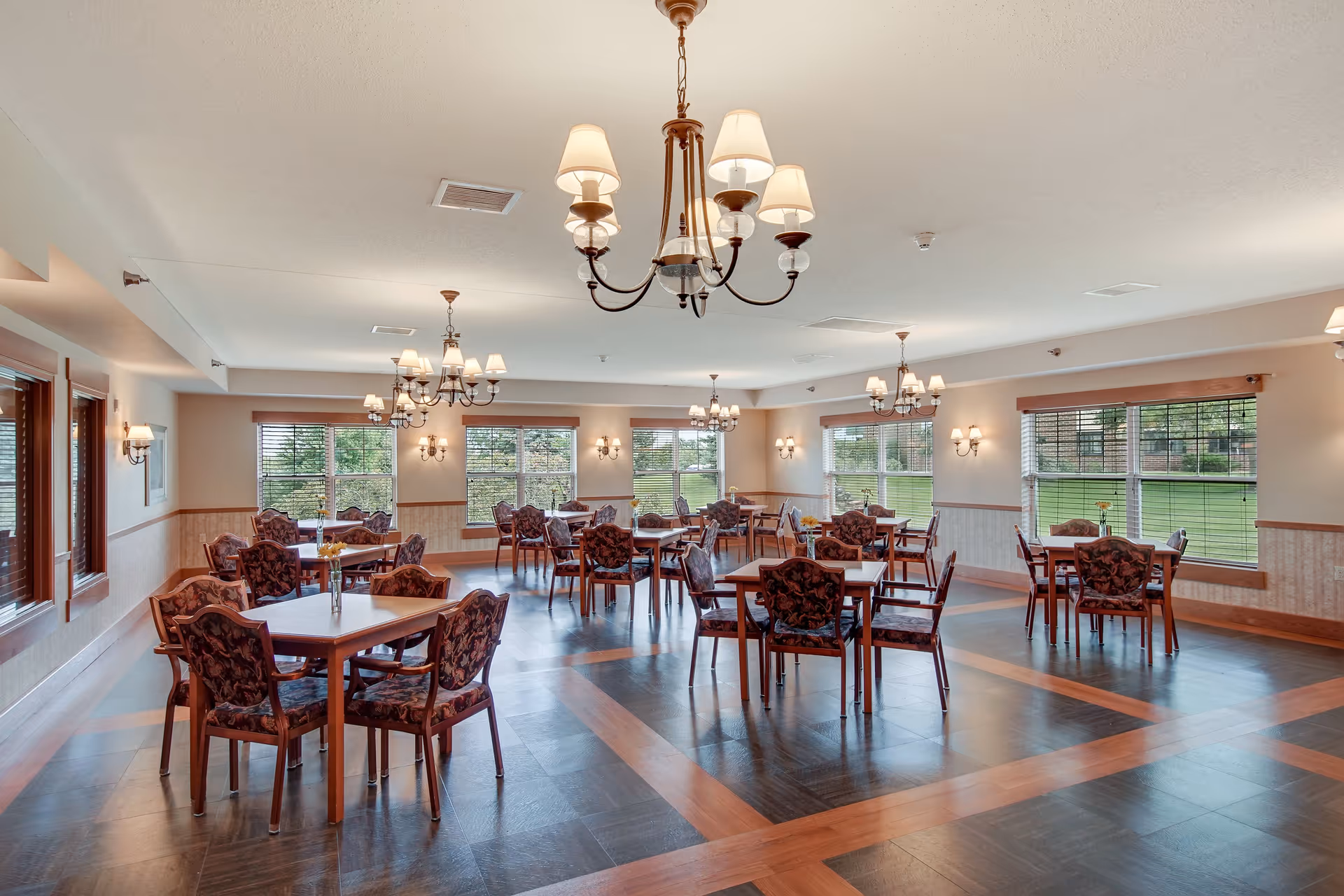A spacious dining room with multiple wooden tables and cushioned chairs arranged neatly. The room features large windows allowing natural light to brighten the space, elegant chandeliers hanging from the ceiling, and wall sconces providing additional lighting. The floor has a patterned design with wood and dark tiles.