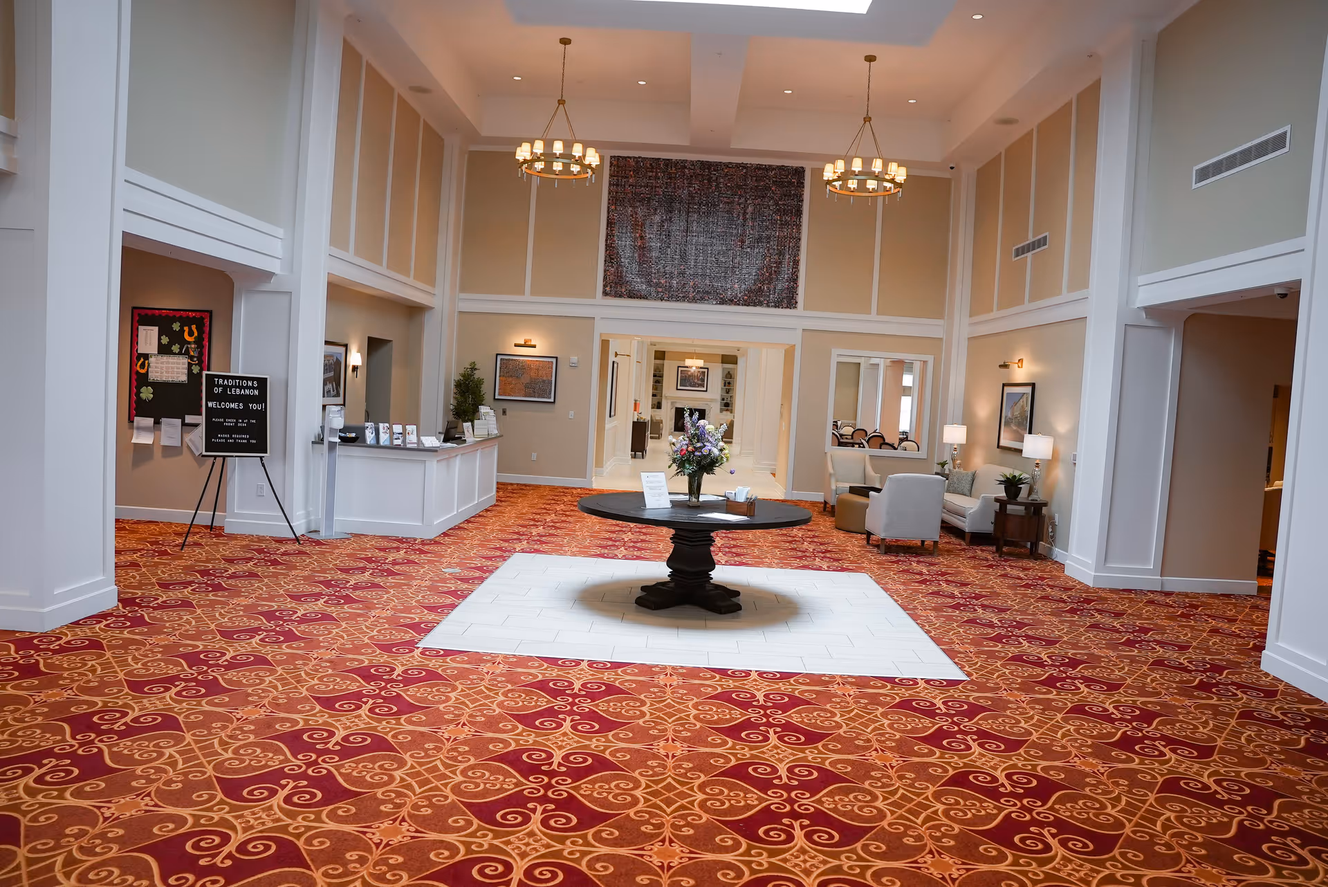 Spacious and elegant lobby area of Traditions of Lebanon featuring a patterned red and orange carpet, a round black table with a flower arrangement in the center, beige walls with white trim, two chandeliers hanging from the ceiling, a reception desk on the left, and a seating area with chairs and lamps on the right.