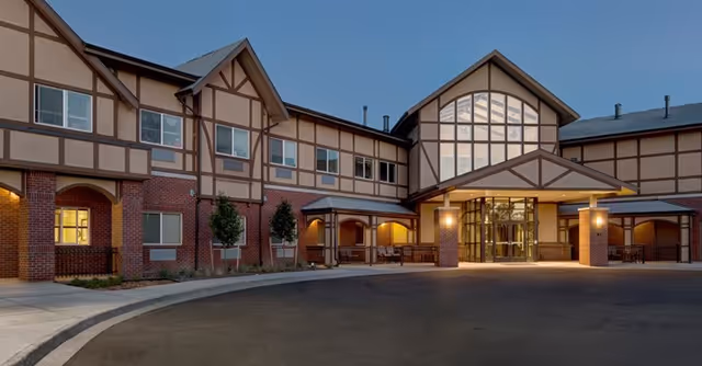 Exterior view of a two-story senior living facility building with Tudor-style architecture, featuring brick and beige paneling, large windows, and a covered entrance with warm lighting at dusk.